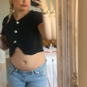 Svart croppad topp med knappar - Svart croppad topp med kort ärm och snygga stora silvriga knappar framtill. Toppen har en lätt v-ringad design och är figurnära. Perfekt till jeans eller kjol för en trendig look.