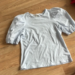 Ljusblå topp med puffärm från H&M - Supersöt ljusblå topp från H&M med puffiga korta ärmar och rund halsringning. Toppen har en clean look och är gjord i mjuk bomull med ärmar i ett lite glansigare material. Perfekt för dig som gillar en feminin och trendig stil.