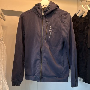 Mörkblå hoodie från Sail Racing - Mörkblå hoodie från Sail Racing med dragkedja hela vägen, huva med snörning och en bröstficka med logga. 