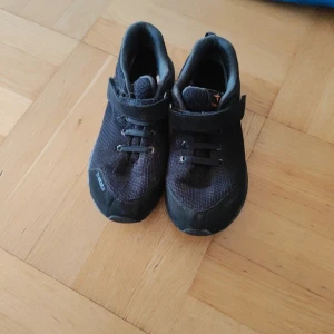 Svarta sneakers från Treksta Comfort-Fit - Svarta sneakers från Treksta Comfort-Fit med mesh-ovandel och praktisk kardborreknäppning. Skorna har platt sula, rund tå och detaljer i syntetmaterial. Perfekta för dig som vill ha bekväma och stilrena skor till vardagen.