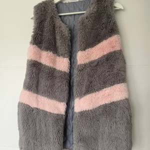 Grå och rosa fluffig pälsväst - Supermjuk och lång pälsväst i grått med breda, snedställda rosa ränder. Västen har quiltat foder på insidan och praktiska fickor. Perfekt för dig som vill ha en mysig och trendig look med extra edge.