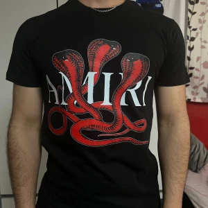 Svart Amiri t-shirt med röd orm - Svart t-shirt från Amiri med stor, röd orm-grafik och vit logga framtill. Klassisk passform med korta ärmar och rund hals. Grym statement-piece för dig som gillar streetwear och coola prints.
