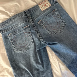 True Religion ljusblå bootcut jeans - Ljusblå jeans från True Religion med ikoniska bakfickor och rosa kontrastsömmar. Modellen har bootcut-ben.  Klassisk femficksdesign och knappgylf . Tyvärr gått sönder lite där nere men inget som åker upp mer eller så, blir finare att klippa av biten som hänger löst där nere men har inte gjort det än ❤️ Säljer då jag tröttnat och dem kommer inte till användning 