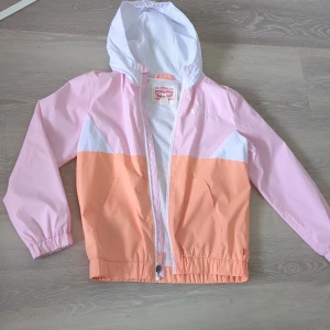 Pastellfärgad vindjacka från Levi's - Snygg vindjacka från Levi's i pastellrosa, persikofärgad och vit. Jackan har huva, dragkedja framtill och två fickor. Tillverkad i lätt polyester med meshfoder för extra komfort. Perfekt för vårens och sommarens äventyr.