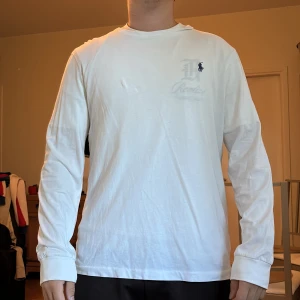 Vit långärmad tröja från Polo Ralph Lauren - Vit långärmad tröja från Polo Ralph Lauren med klassisk blå broderad logga på bröstet. Tröjan har en avslappnad passform och är tillverkad i mjuk bomull, perfekt för en clean och stilren look.