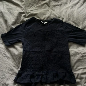 Mörkblå blus med volangkant - Snygg mörkblå blus med korta ärmar och smockad midja. Toppen har en volangkant längst ner som ger en extra touch. Perfekt att styla med jeans eller kjol för en trendig look.