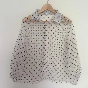 Prickig Blus - Säljer en vit transparent blus med svarta prickar och klassisk krage. Skjortan har långa ärmar med lätt puff och knäppning framtill. Perfekt för dig som gillar trendiga och lekfulla detaljer. Materialet är tunt och ger en luftig känsla. Den är liten i storleken. 