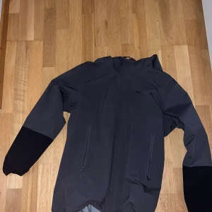 Svart vindjacka från Peak Performance - Snygg svart vindjacka från Peak Performance med huva och hel dragkedja. Jackan har två praktiska fickor med dragkedja och är tillverkad i ett slitstarkt syntetmaterial. Perfekt för blåsiga dagar och outdoor-äventyr.