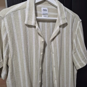 Zara skjorta  - Säljer en beige och vit randig skjorta från Zara i relaxed fit. Skjortan har korta ärmar, klassisk krage och knäppning framtill. Materialet är grovt vävt och ger en chill vibe, perfekt för varmare dagar. Passar dig som gillar en avslappnad och stilren look. Det är storlek L men sitting tight så passar mer M 