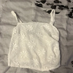 Vit blommig virkad linnetopp - Superfin vit linnetopp med virkade blommor och breda axelband. Toppen har ett genombrutet virkat mönster med broderade blommor över hela och är fodrad på insidan. Perfekt för sommarens alla tillfällen och ger en söt och trendig look.