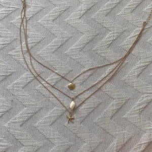 Guldigt halsband med snäckor och sjöstjärna - Säljer ett trendigt lager-på-lager halsband i guldton med tre hängen: en snäcka, en vit pärlformad detalj och en sjöstjärna. Perfekt accessoar för dig som gillar strandvibbar och vill addera lite sommarkänsla till din stil.☀️🐚💕