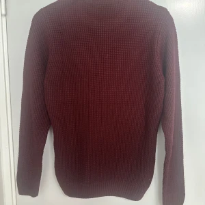 Vinröd stickad tröja från DeFacto - Vinröd stickad tröja från DeFacto med grov struktur och rund hals. Tröjan har långa ärmar och ribbade muddar vid ärmslut och nederkant. Perfekt för dig som gillar en enkel men snygg look under höst och vinter.