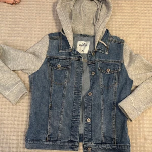Jeansjacka med grå hoodie från Hollister - Snygg jeansjacka från Hollister med grå sweatshirt-ärmar och huva. Klassisk blå denim med två bröstfickor och knappar framtill. Inte använd på över fem år. Inga defekter. Storlek M