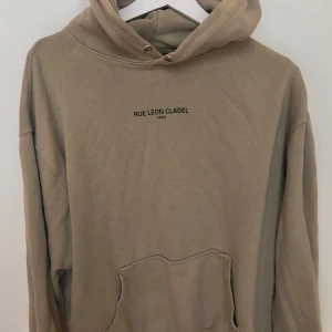 Beige hoodie från Speechless M - Snygg beige hoodie från Speechless med trycket 'RUE LÉON CLADEL PARIS' på bröstet. Klassisk känguruficka framtill, huva med metallöljetter och avslappnad passform. Perfekt för chill dagar och streetstyle.
