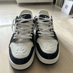 Louis Vuitton svartvita sneakers - Snygga Louis Vuitton sneakers i svart och vitt med tydliga LV-loggor på tungan, hälen och sidan. Skorna har snörning, rund tå och platt sula. Ovandelen är i skinn och textil med monogramdetaljer och kontrasterande paneler. Perfekta för dig som vill sticka ut med lyxig streetstyle.