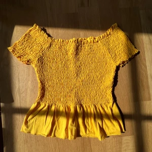 Gina tricot gul topp med volang - Superfin gul offshoulder topp med smockad överdel och volang nertill. Kortärmad och perfekt för soliga dagar. Sitter tajt upptill och har en söt, rynkad effekt. Lätt och luftig känsla.