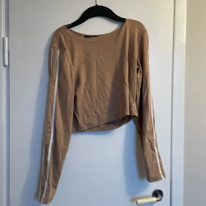 Beige croppad tröja med vita ränder - Säljer en beige croppad tröja med långa ärmar och vita ränder längs ärmarna. Perfekt för en avslappnad stil. Tröjan har en rund halsringning och är gjord i ett mjukt material.