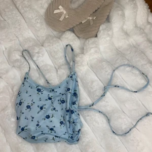 Blå blommig linnetopp - Söt blå linnetopp med blommigt mönster och smala axelband. Toppen har en knytbar detalj på sidan som ger en fin passform. Perfekt för varma sommardagar!