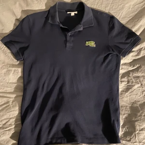 Mörkblå pikétröja från Lacoste - Säljer en klassisk mörkblå pikétröja från Lacoste med korta ärmar och den ikoniska krokodilloggan på bröstet. Perfekt för en stilren och avslappnad look. Passar bra till både jeans och chinos.