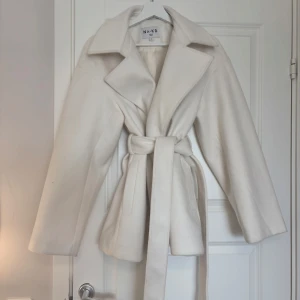 Vit kappa från NA-KD - Elegant vit kappa i storlek S från NA-KD med klassisk krage och knytskärp i midjan. Aldrig anvönd. Stilrena design och långa ärmar. En tidlös favorit i garderoben! Pris 400kr