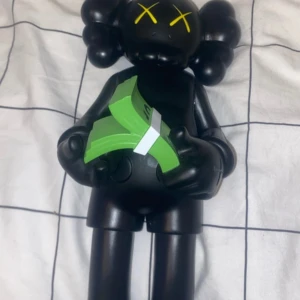 Svart figur med gröna sedlar - Säljer en unik svart figur med karakteristiska korsade ögon i gult. Figuren håller en bunt gröna sedlar i händerna. Perfekt för samlare eller som en cool inredningsdetalj.