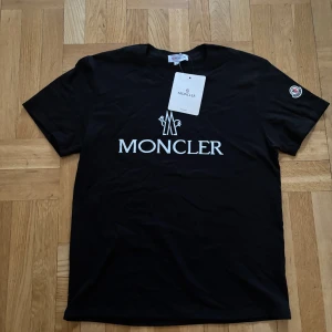 Svart t-shirt från Moncler - Snygg svart t-shirt från Moncler med deras ikoniska logga tryckt på bröstet och en patch på ärmen. Perfekt för en stilren look. T-shirten är kortärmad och gjord i mjukt material för extra komfort. Passar M/L