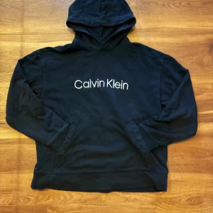 Svart hoodie från Calvin Klein - Säljer en svart hoodie från Calvin Klein med deras logga tryckt på framsidan. Endast använd ett fåtal gånger så är hyfsat ny. Säljer dock för att den inte passar längre. Tröjan har en avslappnad passform och en praktisk magficka. Perfekt för en casual look.