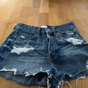 Blå jeansshorts från H&M i stl 36 - Säljer ett par coola blå jeansshorts från H&M med slitna detaljer och fransar. Perfekta för en avslappnad stil. De har en klassisk femficksdesign och knappgylf.