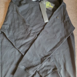 stone island tröja - Stilren svart SI tröja. Tröjan har långa ärmar och en rund halsringning. Den är prydd med det ikoniska märket på ärmen. Helt oanvänd och i storlek M.