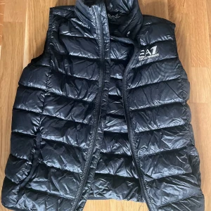 Svart dunväst från Emporio Armani - Snygg svart dunväst från Emporio Armani med dragkedja och hög krage. Perfekt för kyligare dagar och ger en stilren look. Märkeslogga på bröstet för en exklusiv touch.