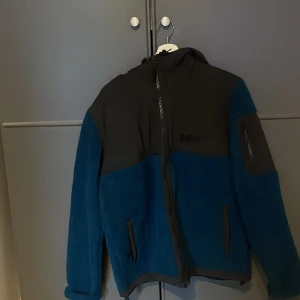 Blå och svart fleecejacka från Helly Hansen - Säljer en snygg blå och svart fleecejacka från Helly Hansen. Jackan har en dragkedja framtill och två praktiska fickor med dragkedja. Perfekt för kyliga dagar och outdoor-äventyr. Ny pris 2100 mitt pris 900