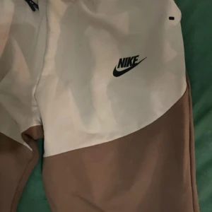 Tvåfärgade mjukisbyxor från Nike - Snygga mjukisbyxor från Nike i en cool tvåfärgad design med beige och brun. Byxorna har en elastisk midja med snörning och Nike-loggan tryckt på benet. Perfekta för en avslappnad stil.