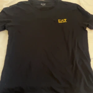 Svart t-shirt från Emporio Armani - Säljer en stilren svart t-shirt från Emporio Armani med gul logga på bröstet. Perfekt för en casual look. T-shirten har en normal passform och är kortärmad.