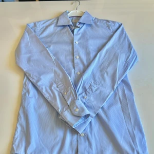 Blå randig skjorta från Eton - Snygg blå skjorta från Eton med smala vita ränder. Skjortan har en slim passform och långa ärmar med knappar. Perfekt för en stilren look.