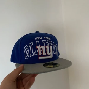Blå New York Giants keps - Snygg blå keps från New Era med New York Giants logga framtill. Kepsen har en grå skärm och är en del av 59FIFTY-serien. Perfekt för sportfantaster och streetwear-entusiaster. Aldrig använd