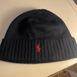 Mörkblå stickad mössa från Ralph Lauren - Snygg mörkblå stickad mössa från Ralph Lauren med röd broderad logotyp framtill. Perfekt för kyliga dagar med sin ribbade kant som ger extra värme och stil. Aldrig använd. 100% merinoull. OBS Juniorstorlek.