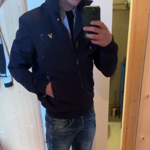  Lyle & Scott - Snygg svart jacka  från Lyle & Scott med gul broderad logga på bröstet. Jackan har dragkedja och ribbade muddar vid ärmslut och nederkant. Perfekt för en stilren look. Det är en liten defekt i armhålan där några trådar har låsnat men det är inget hål och inget som påverkar jackan.