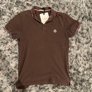 Brun pikétröja från Moncler - Säljer en snygg brun pikétröja från Moncler med korta ärmar och klassisk krage. Tröjan har en broderad Moncler-logga på bröstet och detaljer i rött, vitt och blått på kragen och ärmsluten. Perfekt för en stilren look! 🟢såklart äkta🟢