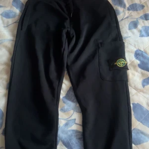 Svarta cargobyxor från Stone Island - Säljer ett par svarta cargobyxor från Stone Island med en praktisk ficka på sidan. Byxorna har en normal passform och är perfekta för en avslappnad stil. Märkets ikoniska logga syns på fickan.