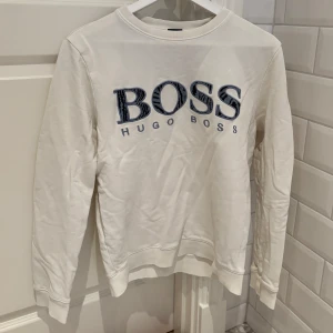 Vit sweatshirt från Hugo Boss - Säljer en stilren vit sweatshirt från Hugo Boss med deras ikoniska logga i blått på bröstet. Tröjan har långa ärmar och en rund halsringning. Perfekt för en avslappnad och trendig look. Något missfärgd på ena axeln men annars bra skick. Nypris: cirka 1000kr 