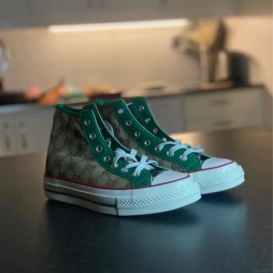 Converse All Star x Gucci sneakers - Unika Converse All Star sneakers i samarbete med Gucci. Skorna har ett brunt monogrammönster med gröna detaljer och vit sula. Klassisk hög modell med snörning och rund tå. Perfekt för den som vill sticka ut med stil.
