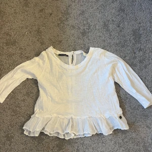 Vit blus med volangdetaljer  XS - Säljer en söt vit blus med volangdetaljer i nederkant och knappar på baksidan. Blusen har trekvartsärmar. Perfekt för en stilren look! En knapp saknas bak i storlek XS