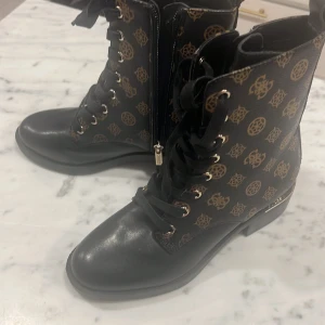 Svarta kängor med mönster och sneakers - Säljer ett par svarta kängor med snörning och dragkedja, dekorerade med ett guldmönster. De har en rund tå och är tillverkade i syntetmaterial. Inkluderar även ett par svarta sneakers med liknande mönster och gulddetaljer.Märke Guess Aldrig använda helt nya  