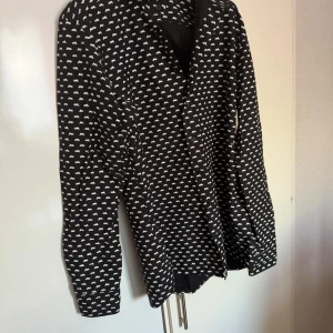 Style of Norrland  - Snygg svart skjorta från Style of norrland med ett coolt vitt mönster. Skjortan har långa ärmar och knappar framtill. Perfekt för att ge din outfit en unik touch.