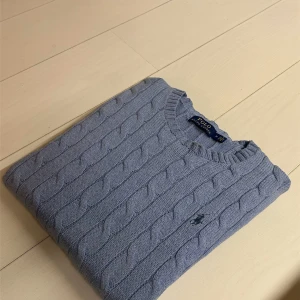 Light blue Ralph Lauren cable-knit sweater - Säljer en stickad tröja från Polo Ralph Lauren med klassiskt flätmönster. Tröjan har en rund halsringning och den ikoniska logotypen broderad på bröstet. Perfekt för en elegant och avslappnad look. Storlek M