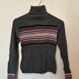 Grå polotröja från KappAhl - Säljer en stilren grå polotröja från Kappahl med randigt mönster i rosa och svart. Tröjan är ribbad och har långa ärmar.