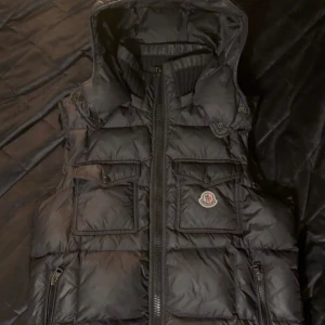 Moncler väst - Snygg svart dunväst från Moncler med huva och dragkedja. Västen har två fickor med knappar på bröstet och en broderad Moncler-logga. Perfekt för kyliga dagar.