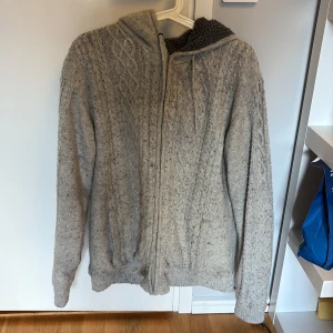 Grå stickad hoodie med dragkedja - Säljer en mysig grå stickad hoodie med dragkedja och huva. Perfekt för kyliga dagar med sin varma och bekväma design. Den har långa ärmar och ribbade muddar för extra komfort.