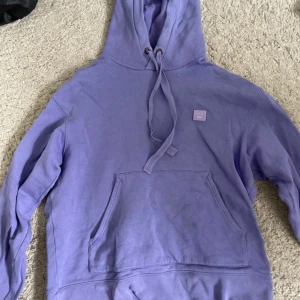 Lila hoodie från Acne Studios - Säljer en snygg lila hoodie från Acne Studios med en klassisk känguruficka och justerbar huva. Den har en liten logotyp på bröstet och är perfekt för en avslappnad stil. Passar bra till både jeans och joggers.