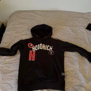 Svart hoodie från Hoodrich - Säljer en svart hoodie från Hoodrich med vita och röda detaljer. Den har en stor logga på framsidan och flera mindre märken. Tröjan har en klassisk känguruficka och justerbar huva med dragsko.